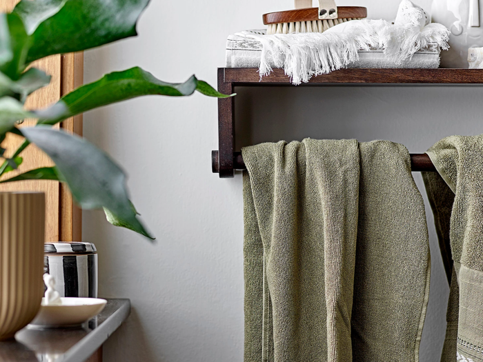 Grønne håndklæder hænger på en håndklædeholder i mørkt træ på et badeværelse med en plante, sæbeskål og toiletartikler i nærheden, hvilket skaber en hyggelig og frisk atmosfære.