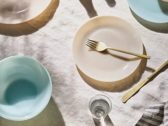 Et solbeskinnet bord med pastelfarvet service, en gaffel på en beige tallerken, et glas vand og en smørkniv på en lys dug.