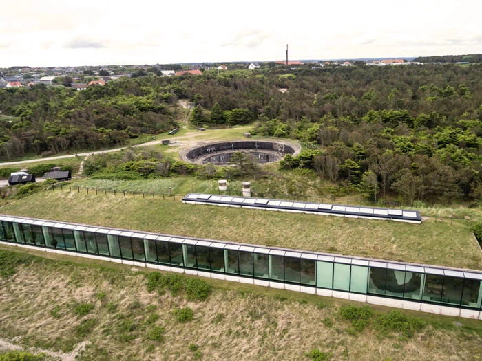 Luftfoto af en moderne bygning med græstag og store glasvinduer ved siden af en cirkulær betonkonstruktion omgivet af træer og grønne områder.
