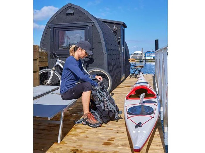 En person sidder på en kaj ved siden af en lille sort hytte, en cykel og en kajak og pakker en rygsæk på en solrig dag nær vandet med både i baggrunden.