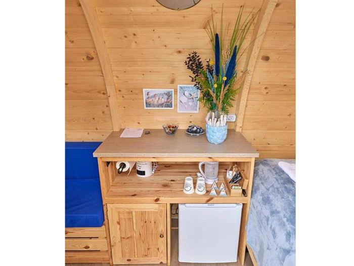 Et lille tekøkken i træ med minikøleskab, krus, kedel og en vase med blomster på en lys væg med træpaneler og kunstværker.