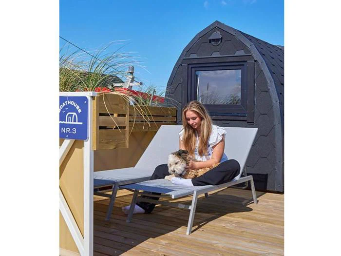 En kvinde sidder i en liggestol på en træterrasse og klapper en lille hund i nærheden af en lille moderne hytte og et skilt med teksten Boathouse NR.3.