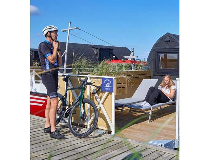 En cyklist i sort tøj står ved siden af sin cykel på et trædæk, mens en kvinde slapper af i en liggestol i nærheden, uden for små moderne hytter.