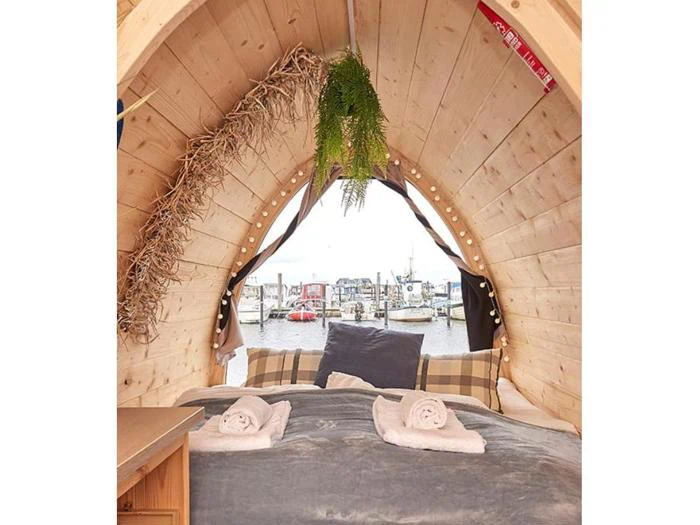 Hyggelig træhytte med en seng, puder og sammenrullede håndklæder, hvor man gennem et buet vindue kan se ud på en marina med både på vandet. Grønne planter hænger ned fra loftet.