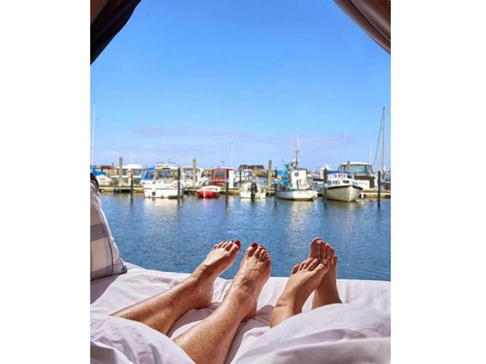 To mennesker ligger i sengen med fødderne oppe og kigger ud på en marina med både, der ligger til kaj under en klar blå himmel.