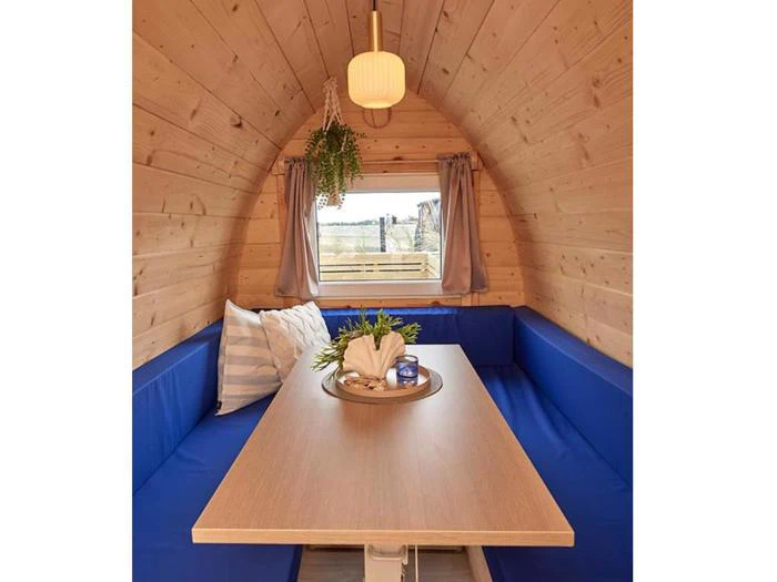 Hyggeligt træhytteinteriør med en blå polstret bænk, et lille bord, en hængende plante, en pendel og et vindue med grå gardiner. En bakke med dekorationer står på bordet.