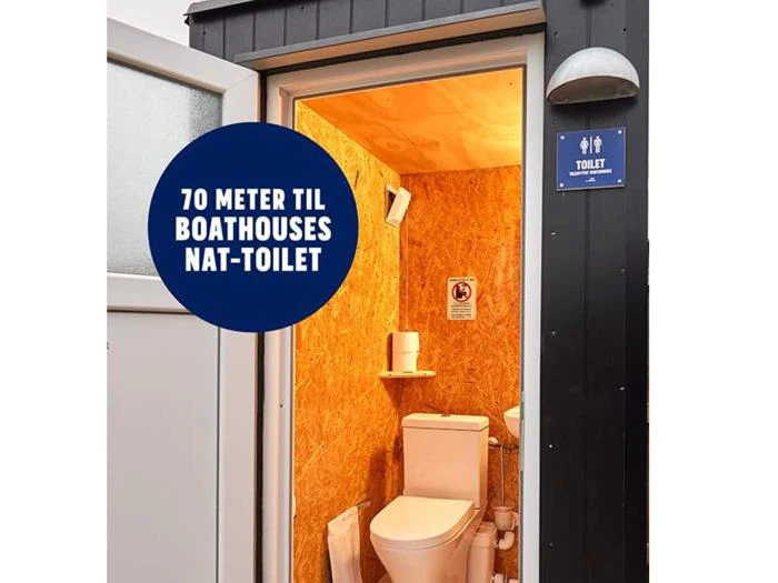 Kig ind i et lille trætoilet med toilet og håndvask; på et skilt over døren står der 70 meter til bådhuse nat-toilet.