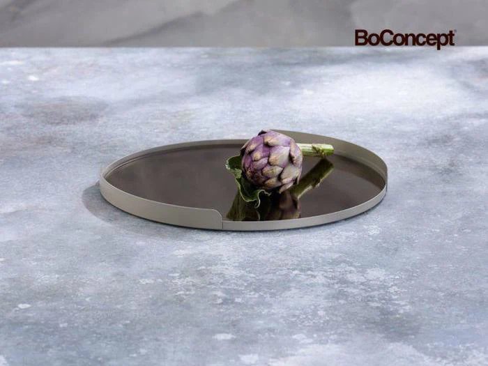 En frisk, lilla artiskok hviler på en rund, mørkegrå BoConcept-bakke, der er placeret på en gråmeleret stenoverflade.