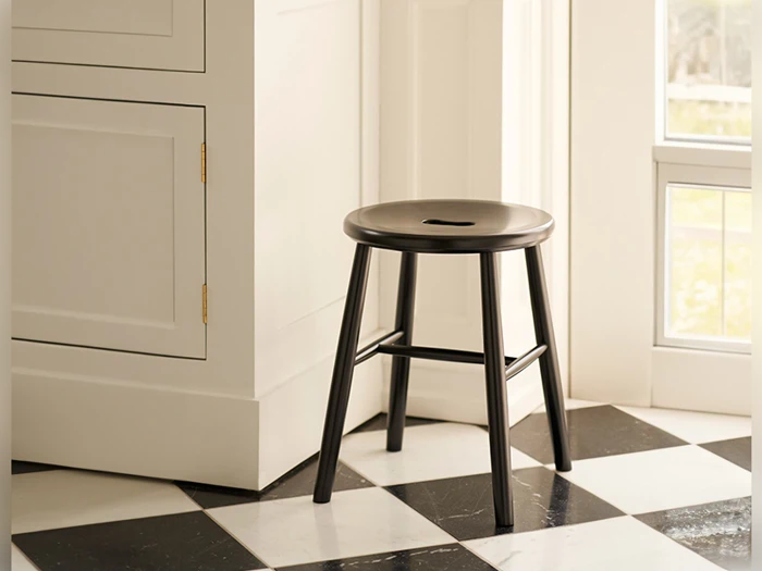 En sort træskammel med en slids i sædet står på et sort-hvidt ternet flisegulv ved siden af et hvidt skab og et vindue.