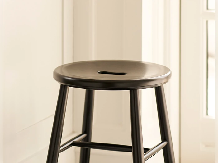 En sort træskammel med et rundt sæde og en udskæring til et håndtag, placeret i nærheden af en hvid væg og et vindue.