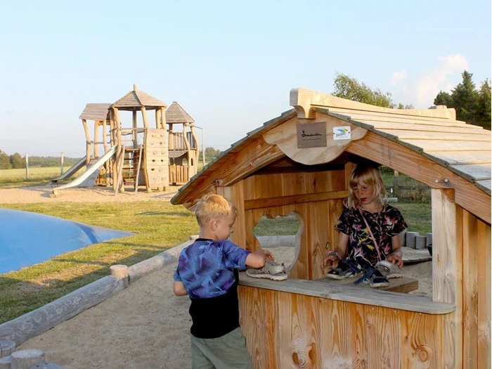 To børn leger med legetøj i et lille legehus af træ på en legeplads med en stor legestruktur af træ og en rutsjebane i baggrunden.