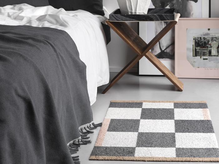 Et sort- og hvidternet tæppe ligger på gulvet ved siden af en seng med et mørkt tæppe; et træstativ og indrammede kunstværker står i nærheden.