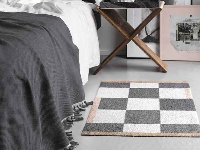 Et ternet tæppe med grå, hvide og beige firkanter ligger på gulvet ved siden af en seng med et mørkegråt tæppe med frynser og en træskammel med en bakke.