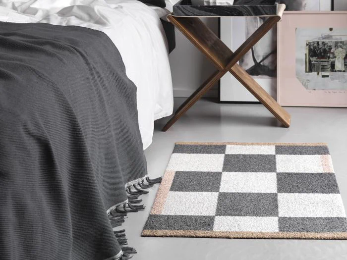 Et ternet tæppe ligger på gulvet ved siden af en seng med et mørkt tæppe og kvaster, i nærheden af et træstativ og indrammede kunstværker.