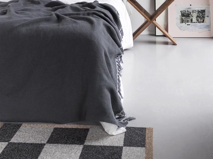 En pænt redt seng med et mørkegråt tæppe, en del af et ternet tæppe og et indrammet kunstværk, der læner sig op ad en hvid væg på et lysegråt gulv.