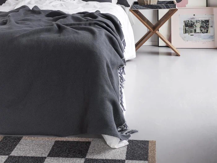 En pænt redt seng med et mørkegråt tæppe, et ternet tæppe, et sidebord i træ og indrammet kunst hviler på et lyst gulv.