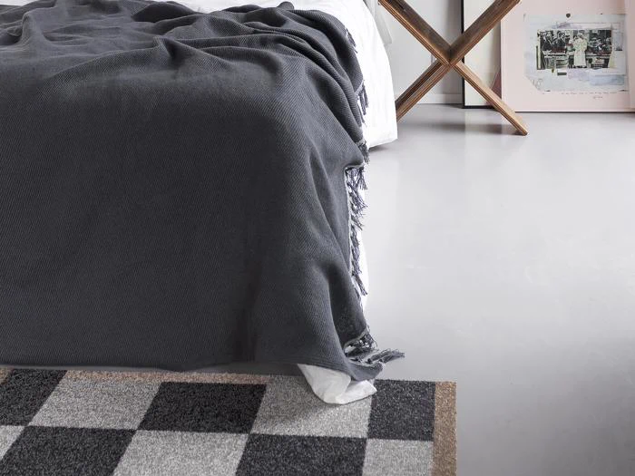 En pænt redt seng med et mørkegråt tæppe, en del af et ternet tæppe og et indrammet kunstværk, der læner sig op ad en hvid væg på et lysegråt gulv.