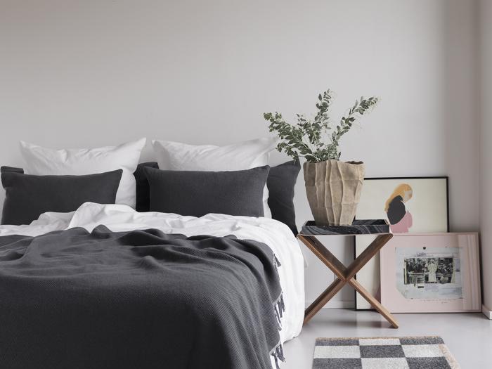 Et moderne, minimalistisk soveværelse med en pænt redt seng med hvidt og mørkegråt sengetøj, et lille sidebord i træ med en potteplante og indrammet kunst, der hviler på gulvet.