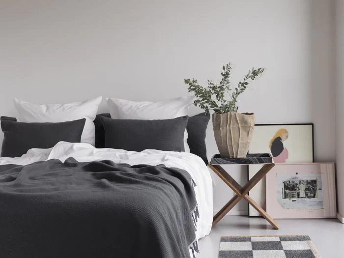 En pænt redt seng med hvide og mørkegrå puder, et gråt tæppe, et lille sidebord i træ med en potteplante og indrammet kunst, der læner sig op ad en hvid væg.
