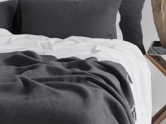 En pænt redt seng med hvide lagner og mørkegråt struktureret sengetøj, inklusive et tæppe og puder, ved siden af et lille sidebord af træ.