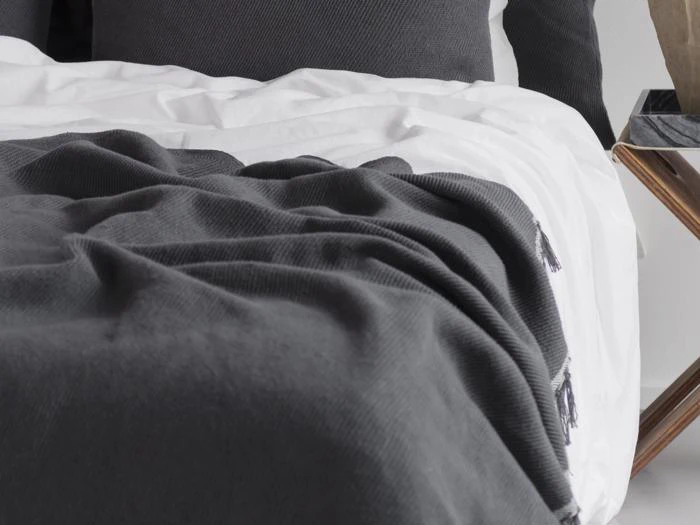 En pænt redt seng med hvide lagner og et mørkegråt struktureret tæppe ved siden af et sidebord af træ.