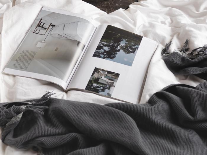 Et åbent magasin med indretningsbilleder ligger på et hvidt lagen ved siden af et mørkegråt tæppe med frynser.