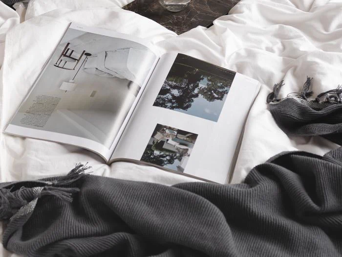 Et opslået magasin ligger på en hvid seng med et mørkegråt tæppe med kvaster i nærheden og viser minimalistiske interiør- og udendørsbilleder.