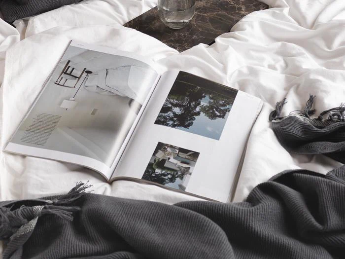 Et opslået magasin ligger på en hvid seng med et sort tæppe og et glas vand i nærheden. Magasinet viser minimalistiske interiør- og udendørsbilleder.