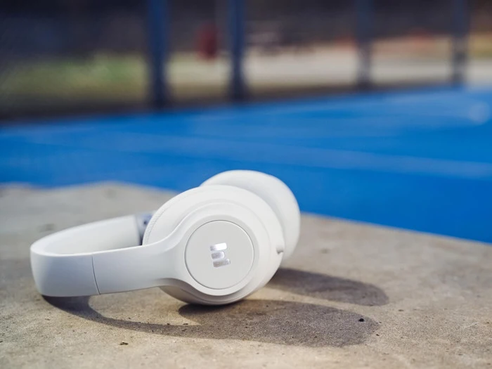Hvide hovedtelefoner hviler på en betonoverflade med en blå udendørs sportsbane og et kædehegn sløret i baggrunden.