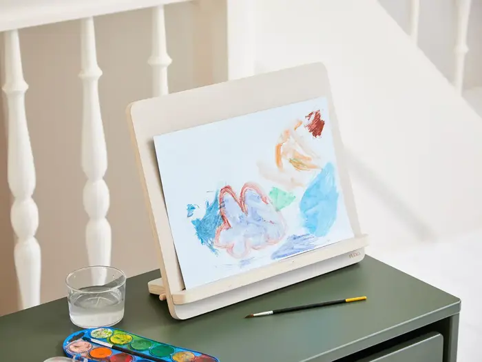 Et barns maleri med farverige abstrakte former står på et træstativ ved siden af en pensel, en vandkop og en akvarelpalet på et grønt bord.