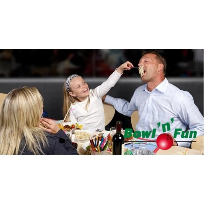 En pige fodrer legende en mand med pasta, mens en kvinde ser på og smiler. De sidder ved et bord med mad på et sted, der hedder Bowl n Fun.
