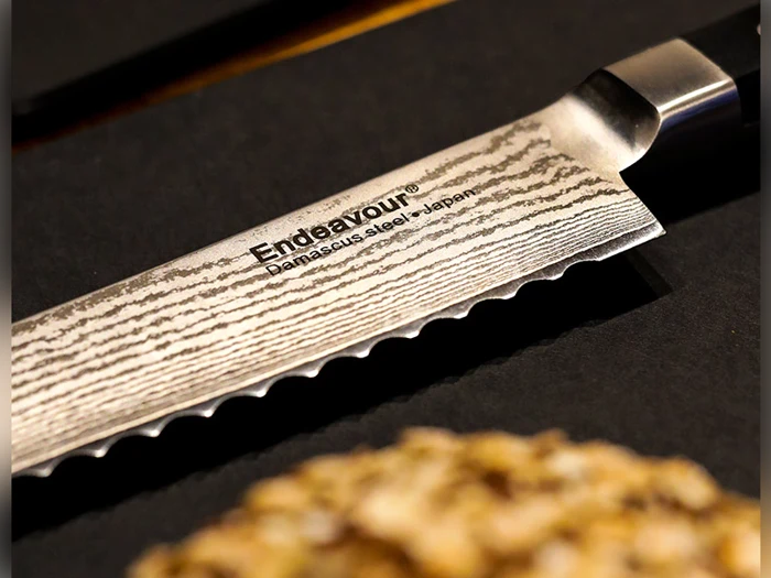 Nærbillede af et savtakket Endeavour-knivblad lavet af damaskusstål med synlige bølgemønstre og teksten Damascus Steel - Japan, placeret på en mørk overflade.