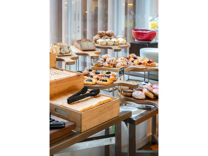 Et buffetbord viser forskellige kager, småkager, brød og donuts på træbakker og stativer, med tallerkener og serveringstænger i nærheden.