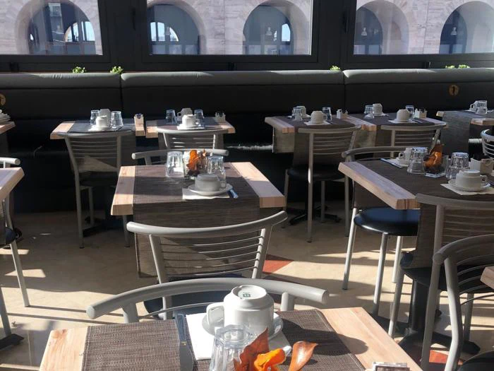 En solbeskinnet restaurant med pænt dækkede borde, hver med kopper, glas, bestik og brune dækkeservietter, der vender ud mod store vinduer med buer udenfor.