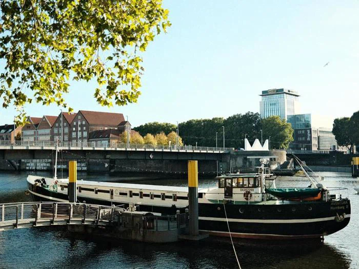 En båd i havn på en flod med en bro, bybygninger og grønne træer i baggrunden under en klar blå himmel.