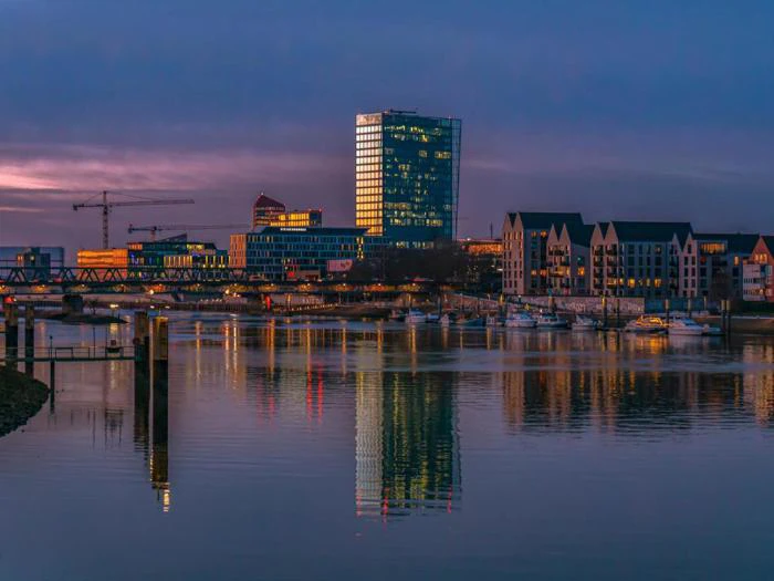 Moderne bygninger og huse ved vandet spejler sig i en rolig flod i skumringen, og byggekraner og både er synlige i billedet.