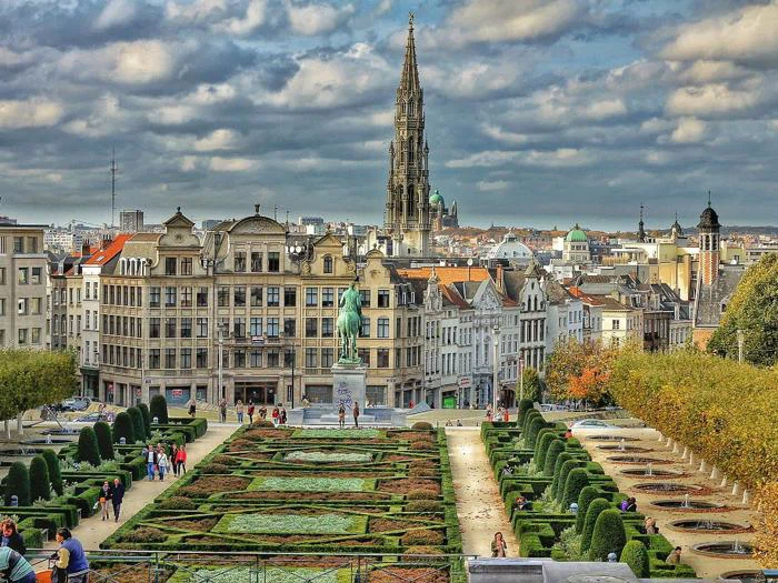 En formel have med klippede hække og stier, omgivet af historiske bygninger og et højt kirkespir under en delvis overskyet himmel i en europæisk by.