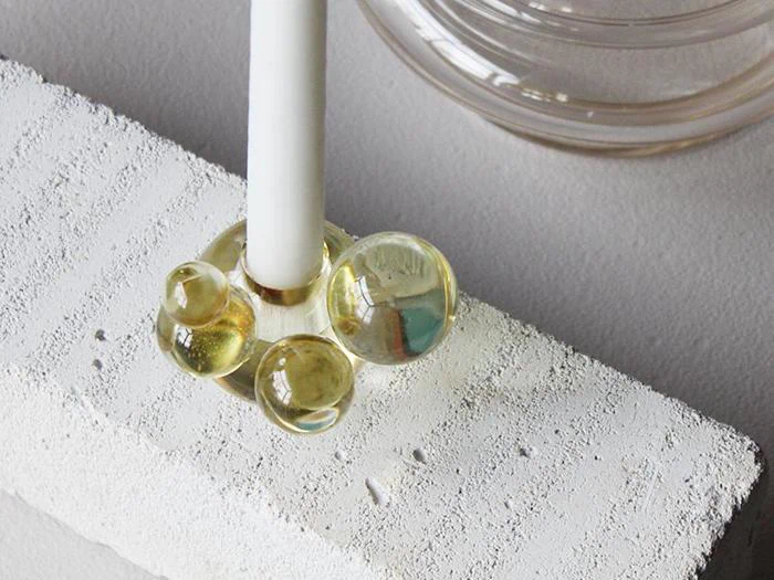 Et hvidt stearinlys holdes oprejst af en glaslysestage med kugleformede former, placeret på en struktureret hvid rektangulær blok.