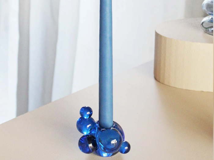 Et højt blåt lys står i en base lavet af klynger af blå glaskugler, placeret på en lys overflade med en blød baggrund.