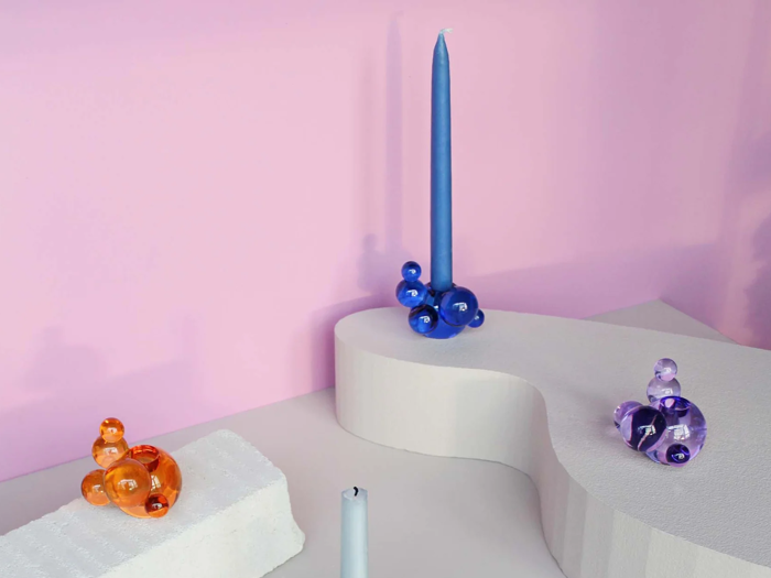 Tre farverige bobleformede lysestager (orange, blå og lilla) med høje stearinlys står på hvide geometriske platforme mod en pink baggrund.