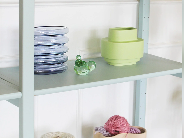 På en lyseblå hylde står stablede klare glastallerkener, en grøn vase, et lille grønt glasornament, en kurv og lyserødt garn. Hvid panelvæg i baggrunden.