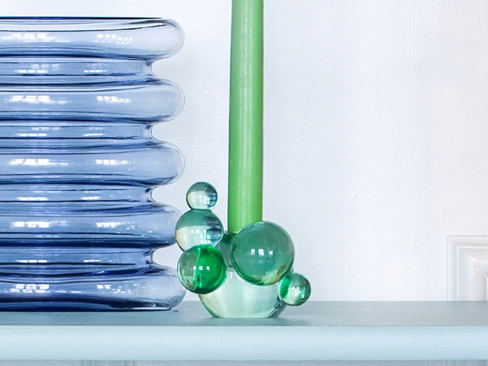 Et grønt stearinlys står i en unik glasholder med grønne og klare glasbobler ved siden af en bølget, gennemskinnelig blå vase på en hvid hylde.