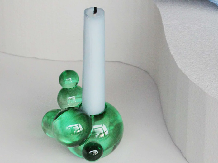 Et hvidt stearinlys står i en blank, grøn glasstage med boblelignende kugler på en hvid, buet baggrund.