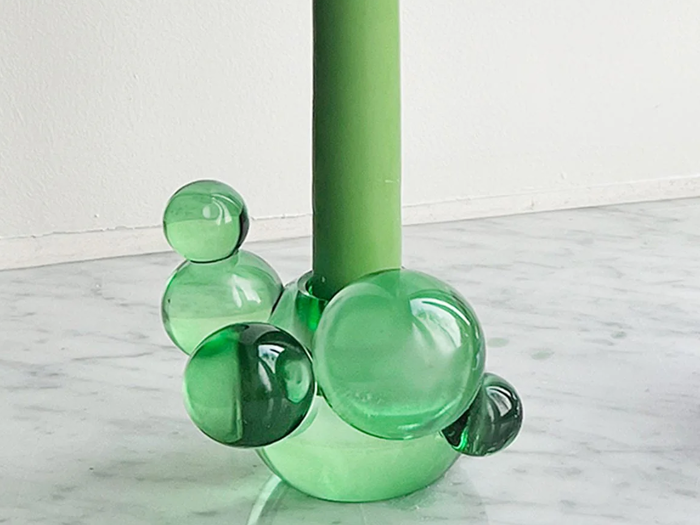 Et grønt stearinlys står i en holder dekoreret med flere gennemsigtige grønne glasbobler på en lys marmoroverflade.