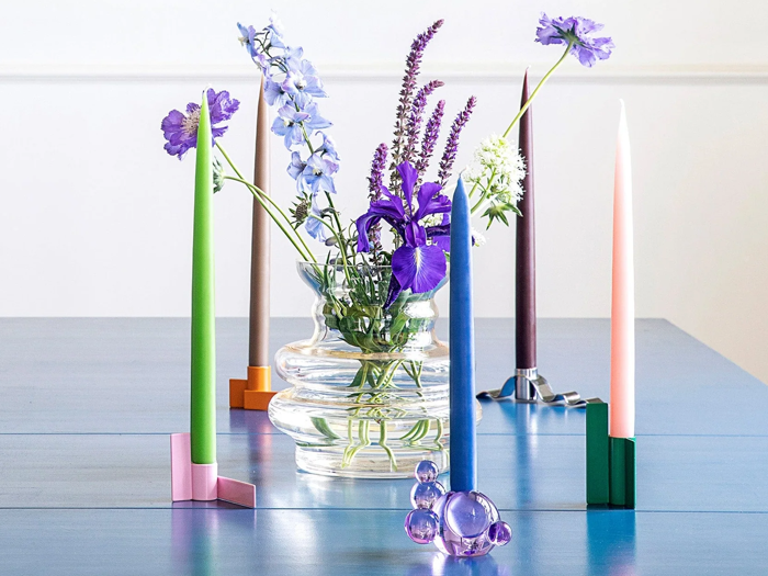 En klar glasvase med lilla og blå blomster står på et blåt bord omgivet af farverige stearinlys i moderne stager.