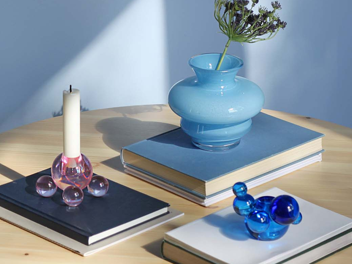 Et rundt træbord med stablede bøger, en blå vase med lavendel, et hvidt lys i en lyserød holder og en lille blå glasskulptur, alt sammen oplyst af blødt sollys.