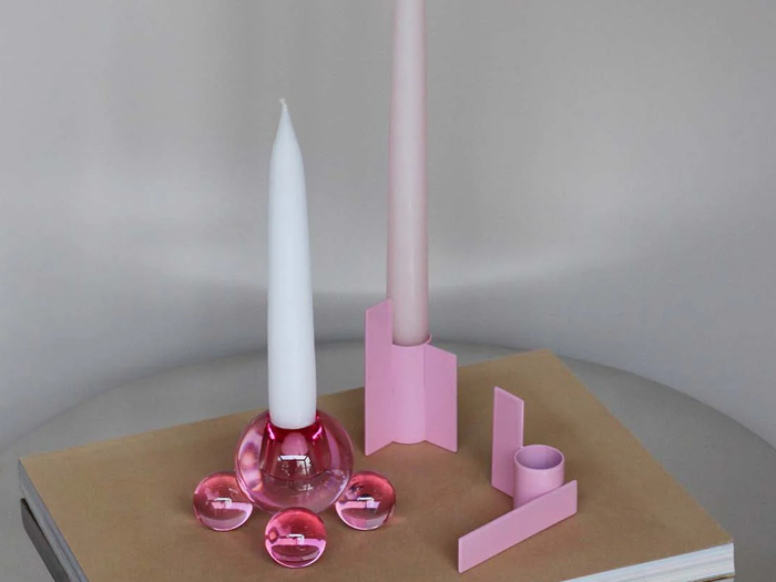 To hvide fyrfadslys i moderne rosa stager på en lukket beige bog med tre små rosa glaskugler i nærheden på en ensfarvet baggrund.