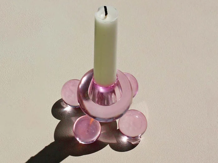 Et hvidt stearinlys står oprejst i en lyserød, gennemsigtig glasstage med cirkulær bund og kaster en blød skygge på en lys overflade.