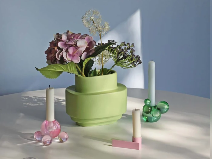 En grøn vase med pink og lilla blomster står på et hvidt bord, omgivet af tre farverige lysestager og pastelfarvede stearinlys, op ad en lyseblå væg.