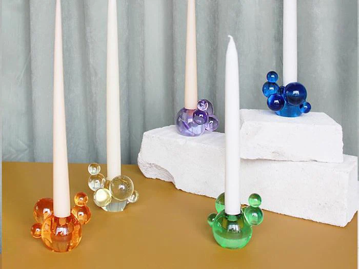 Fem høje lys holdes af farverige, bobleformede glaslysestager i orange, gul, lilla, blå og grøn på en guldfarvet overflade med hvide blokke og en grå baggrund.